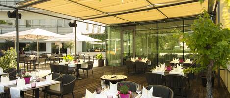 An outdoor dining area with tables and chairs, covered by a canopy, and surrounded by potted plants.