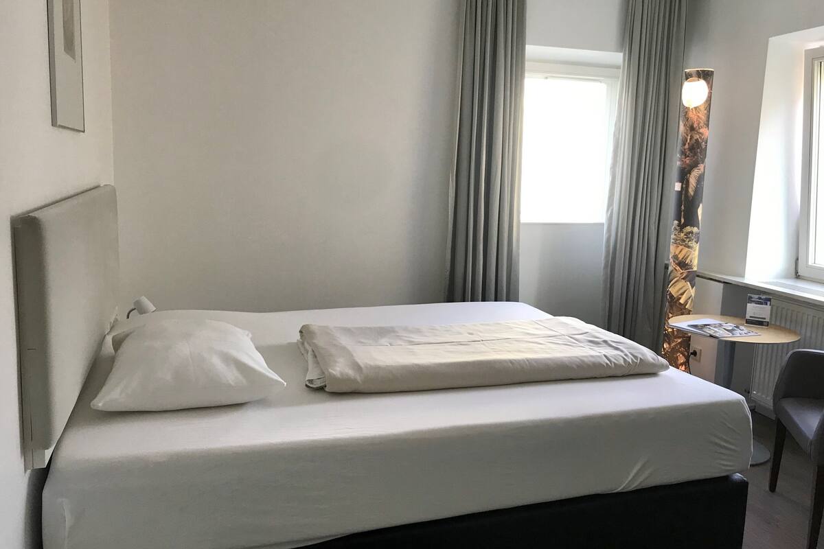 A single bed with white linens, a small bedside table, a chair, and a window with curtains.