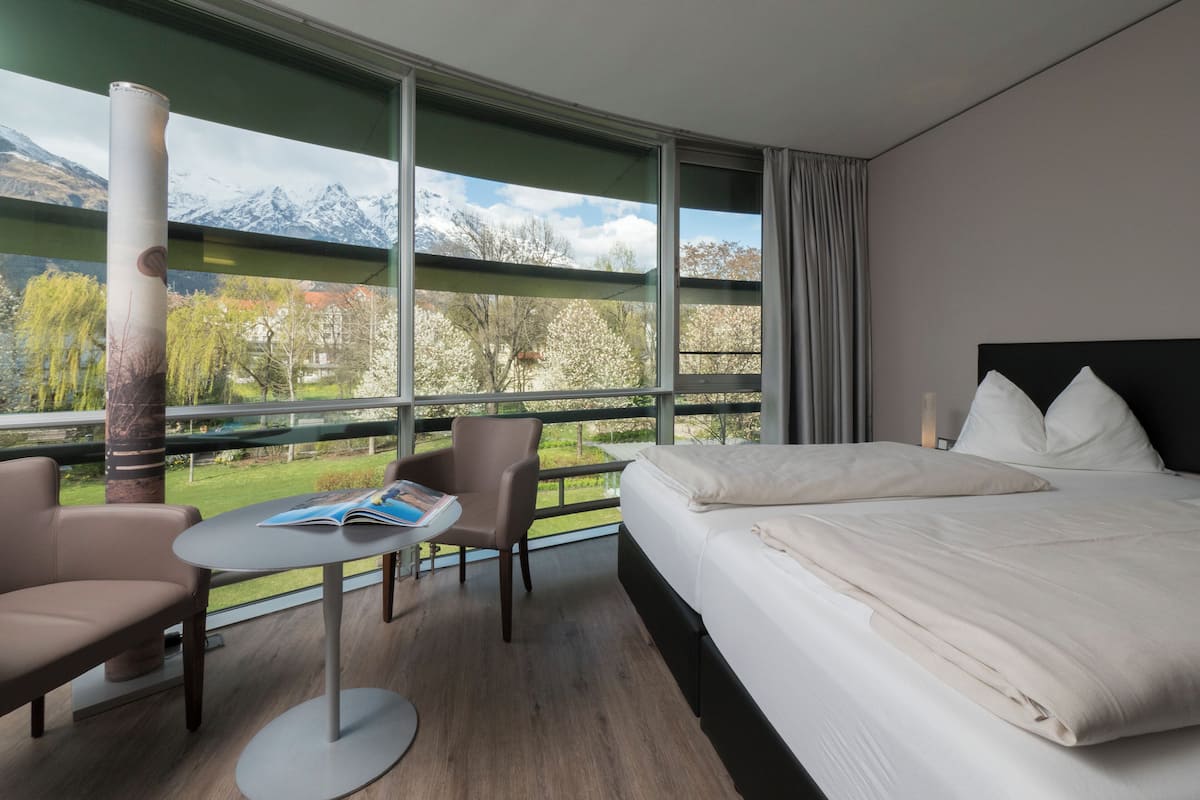 A hotel room with a bed, two chairs, a small table, and a large window offering a mountain view.