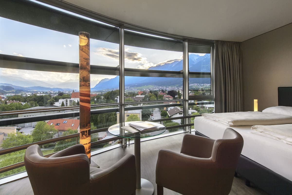 A hotel room with a large window offering a view of a city and mountains. There is a bed, two chairs, and a glass table.