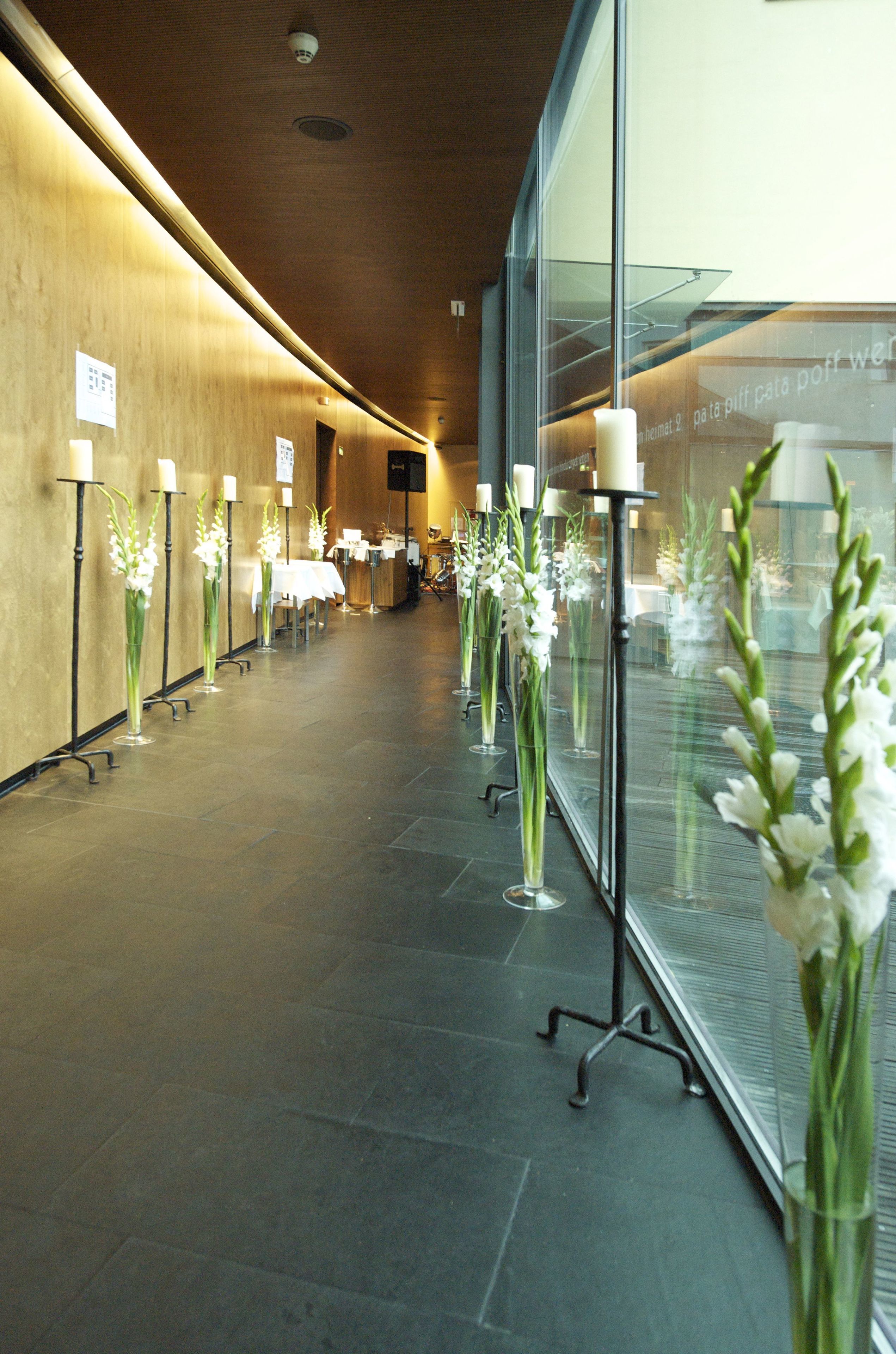 A hallway with a row of tall vases with white flowers, a wooden wall, and a glass partition with a reflection of the room.