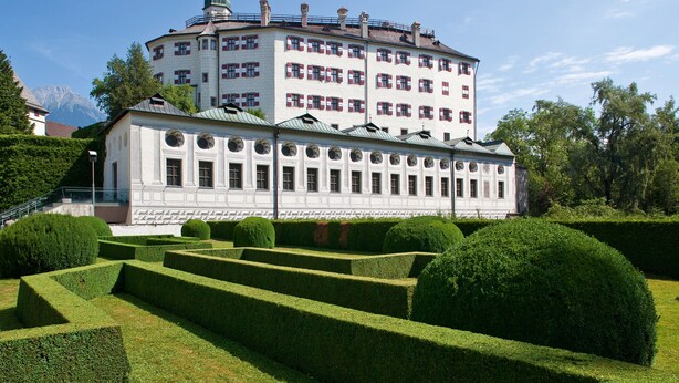 Un edificio grande y blanco con numerosas ventanas y techo verde, rodeado de setos y árboles cuidadosamente podados.