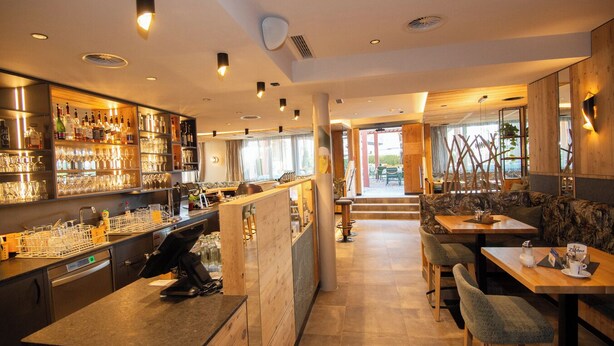 Un bar moderno con mostrador, estanterías llenas de botellas y vasos. Hay mesas y sillas de madera en el comedor, y vistas del exterior a través de amplias ventanas.