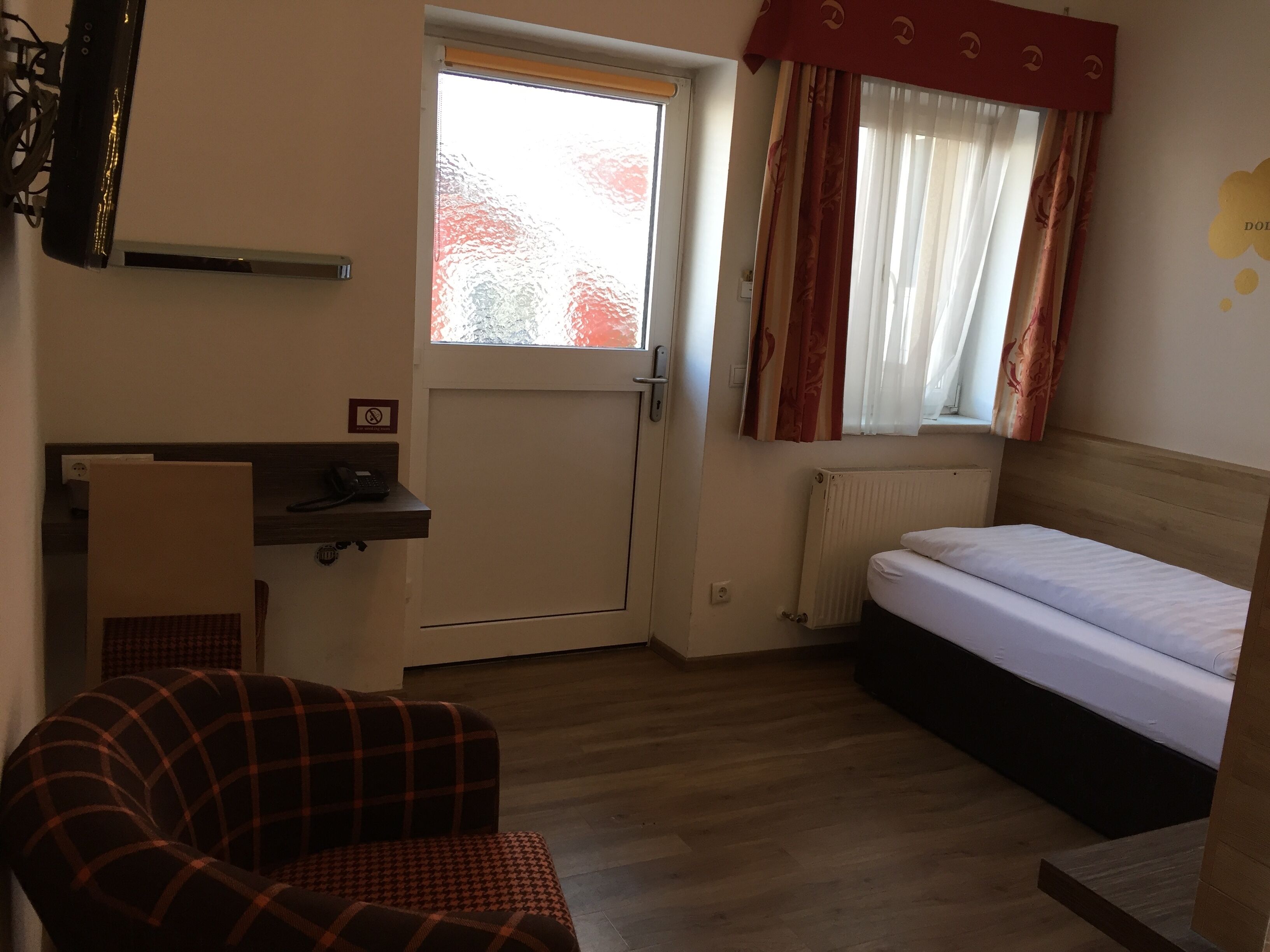 A small, compact room with a bed, a desk with a telephone, a chair, a radiator, and a window with curtains.