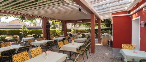 Een buitenpatio met meerdere tafels en stoelen, overdekt door een pergola met glazen panelen.