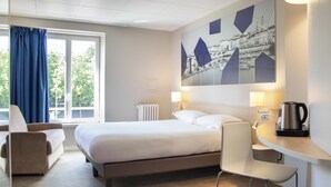 Habitación de hotel con cama, escritorio, silla, radiador y ventana con cortinas.