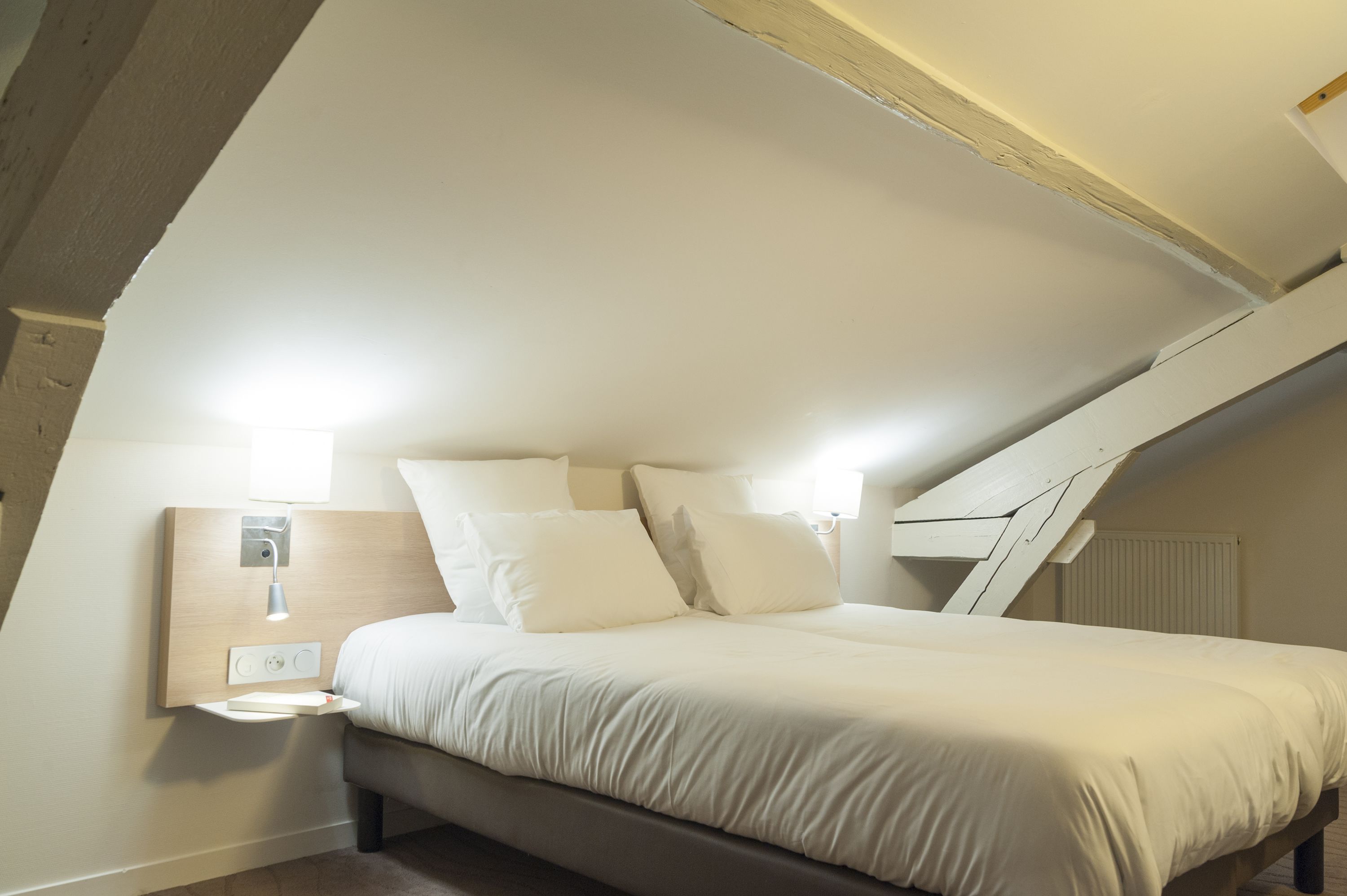 Une chambre avec un plafond en pente, un lit avec une literie blanche, une table de chevet avec une lampe et une prise électrique fixée au mur.
