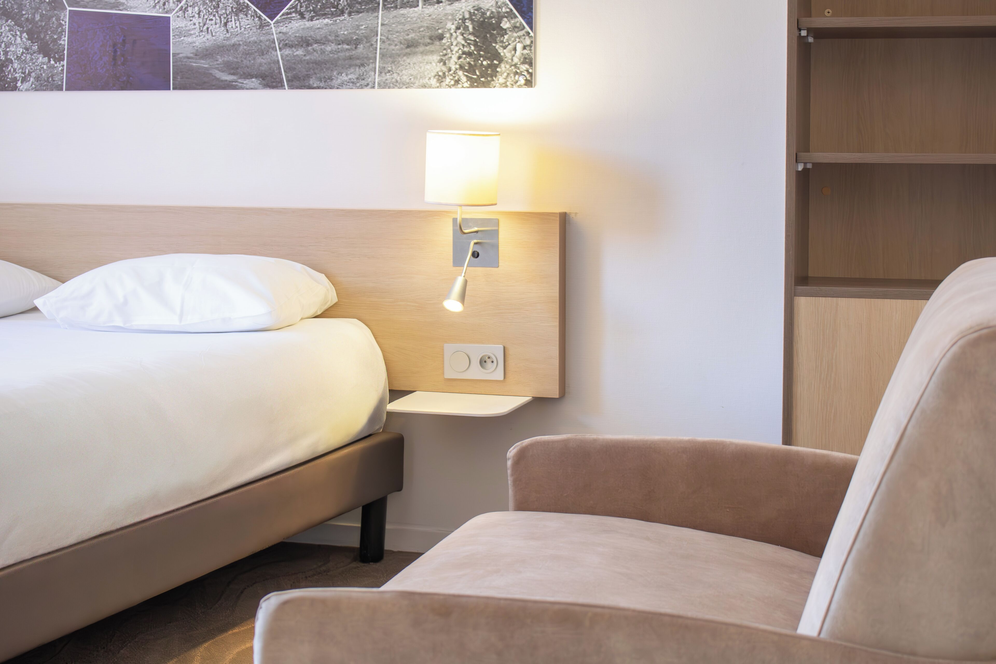 A hotel room with a bed, a bedside table with a lamp, a chair, and a wall-mounted shelf.