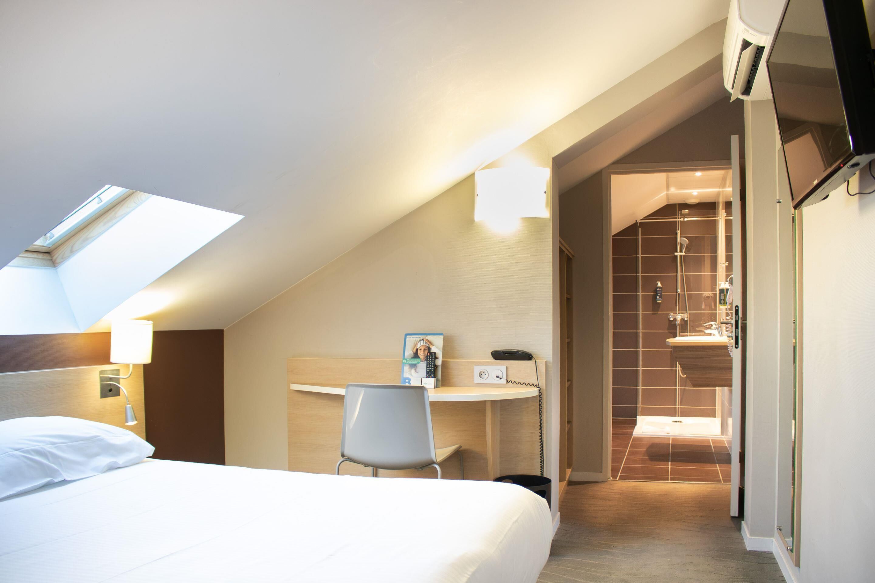 Habitación de hotel con cama, escritorio y silla, teléfono y un marco de fotos. Se observa tragaluz sobre la cama y una zona de ducha a través de una puerta abierta.