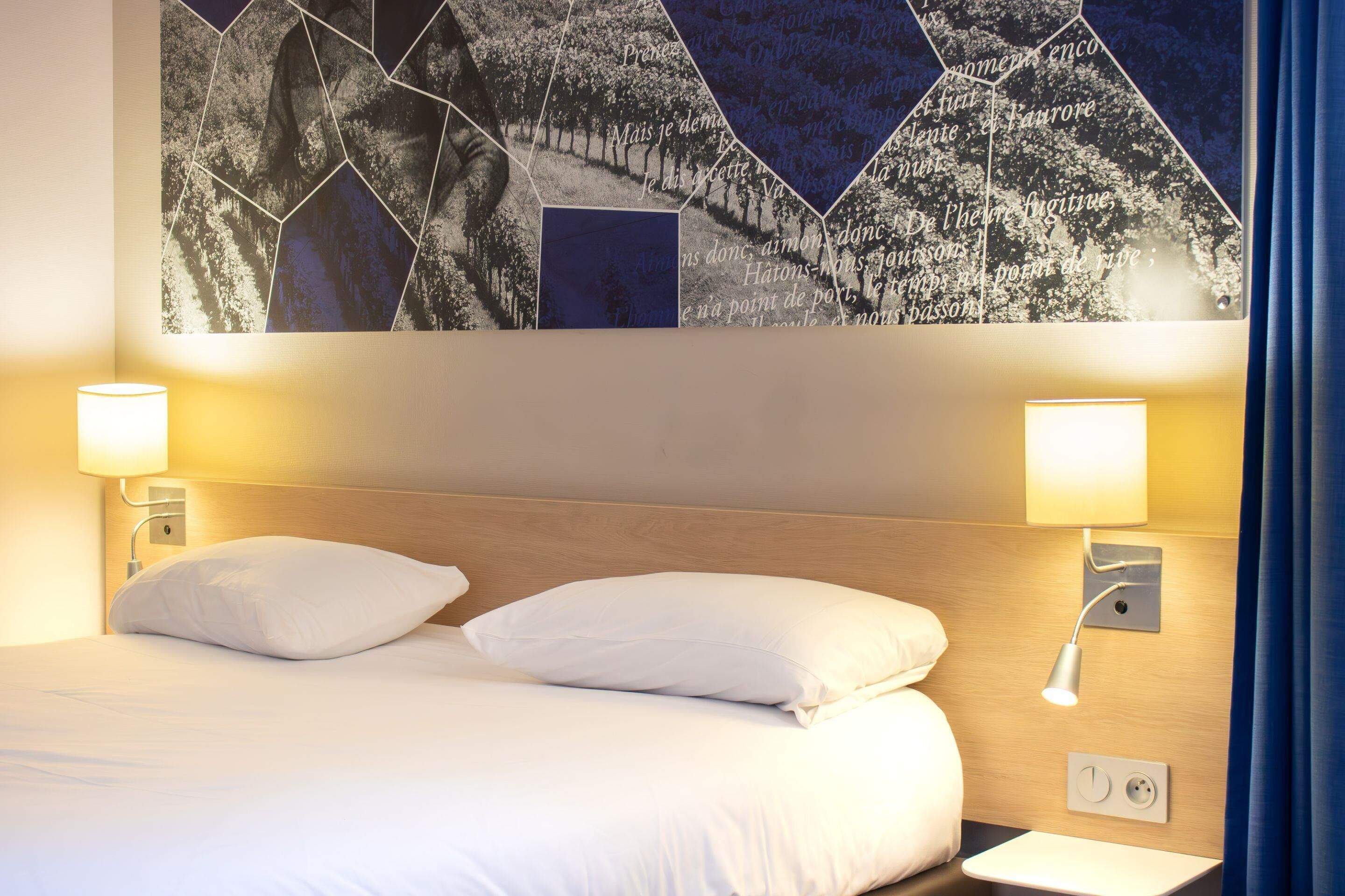 Ein Hotelzimmer mit einem Bett, zwei Nachttischlampen, einer Wandsteckdose und einem großen abstrakten Kunstwerk über dem Bett.