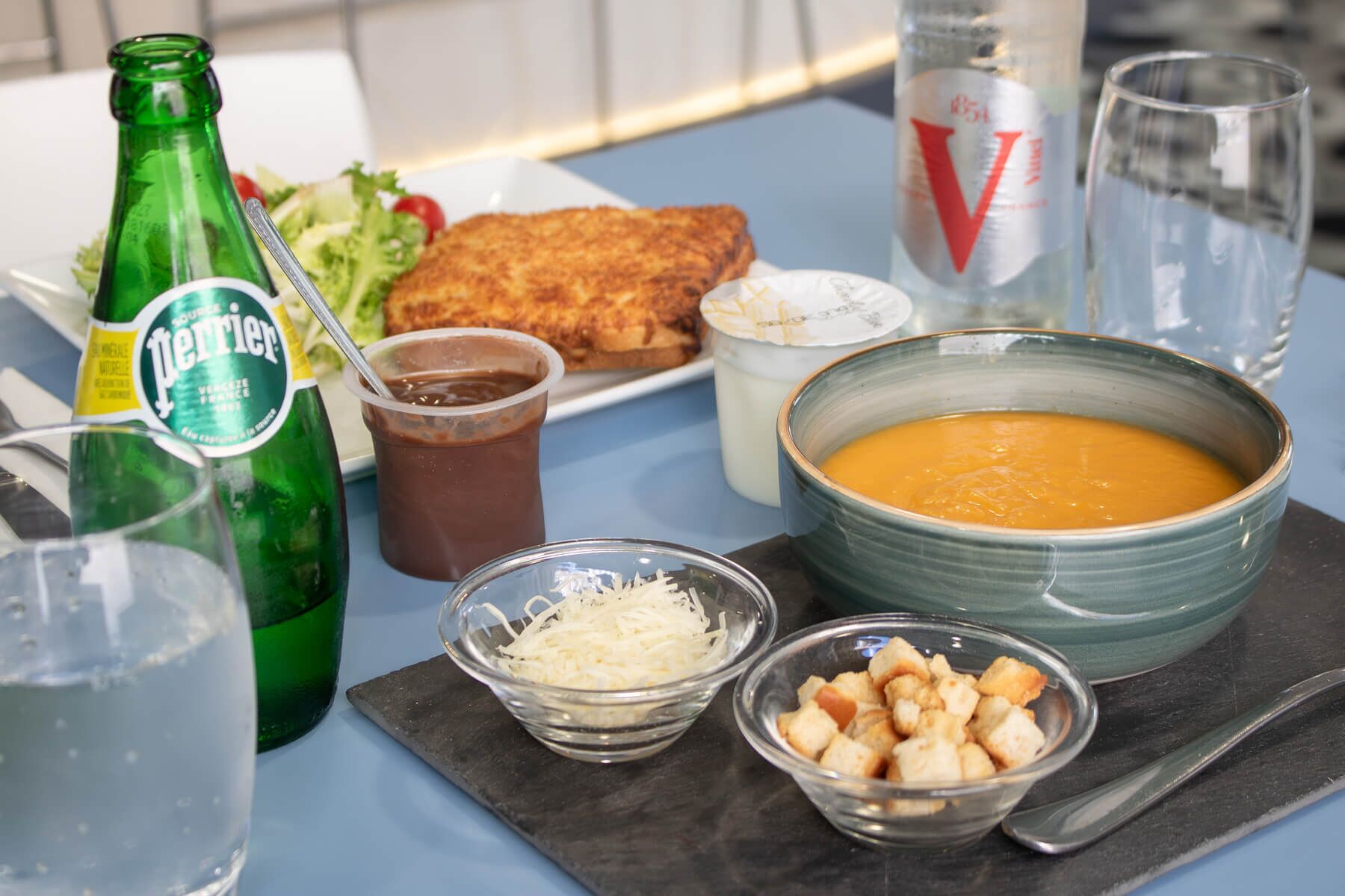 Ett bord med en flaska Perrier, en skål soppa, en tallrik med något friterat, ett glas mjölk och ett glas vatten.