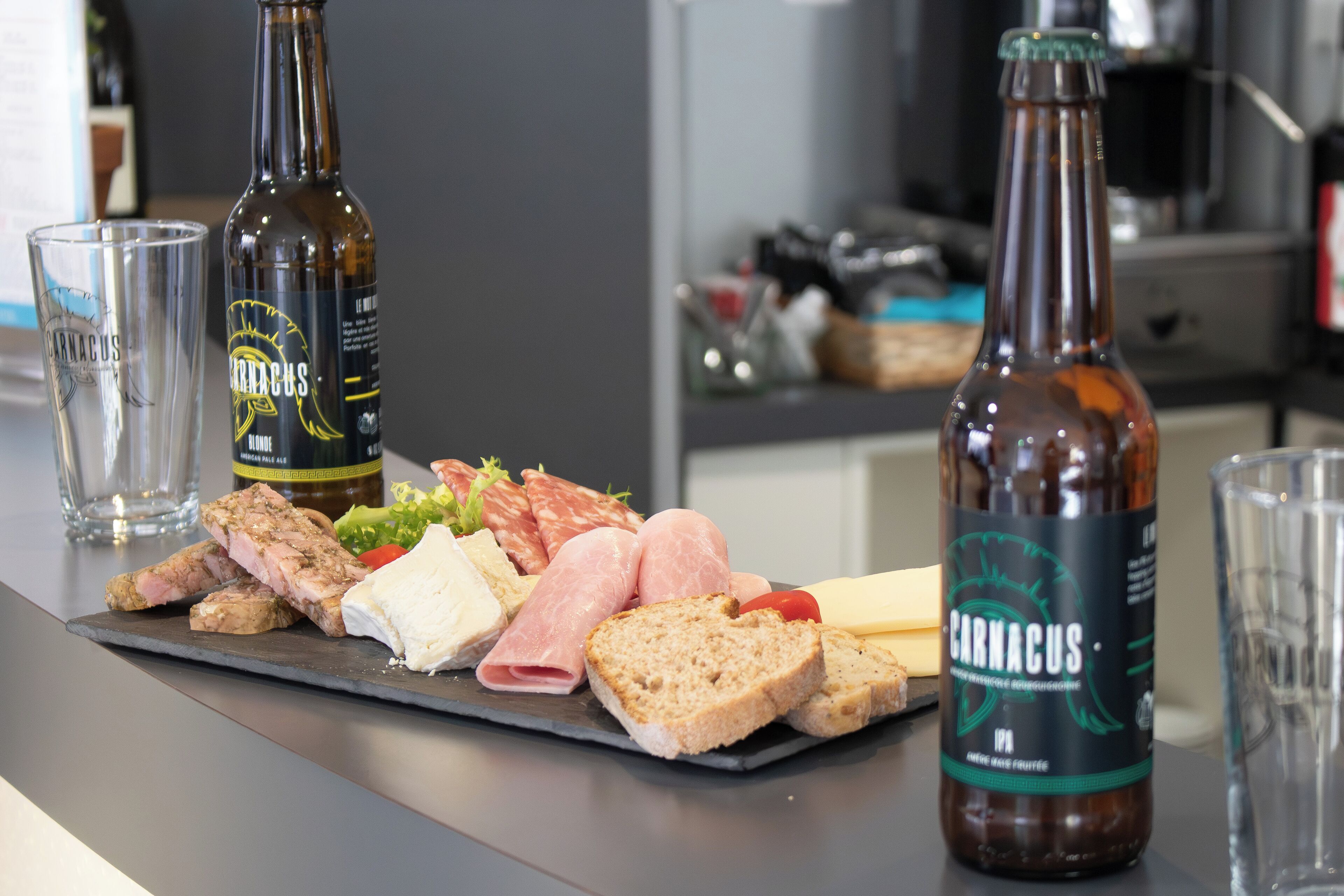 Een charcuterieplank met diverse vleessoorten, kazen en brood, vergezeld van twee flessen Carminius bier en twee glazen op een keukentafel.