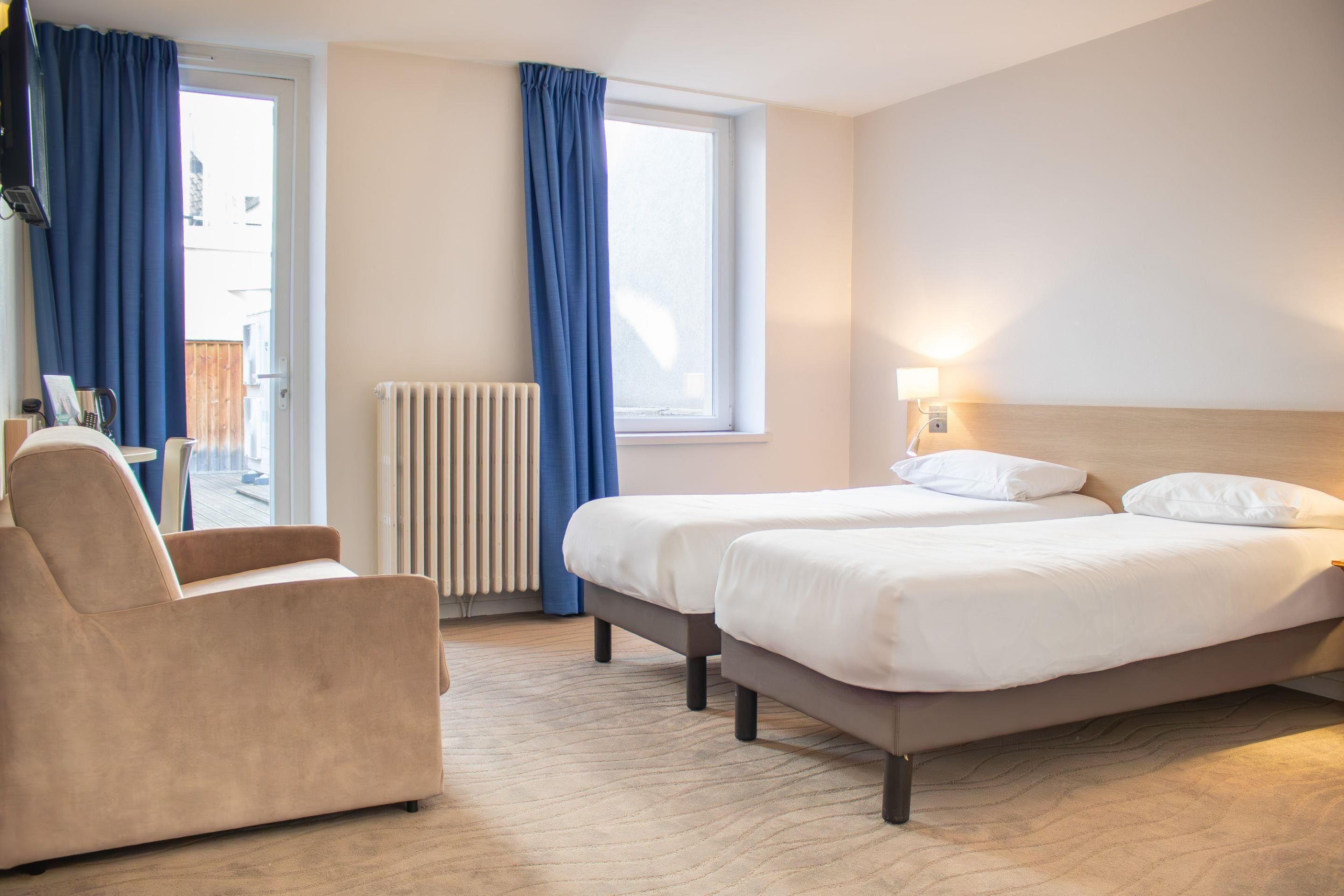 A hotel room with two beds, a chair, a window with curtains, a radiator, and a lamp.