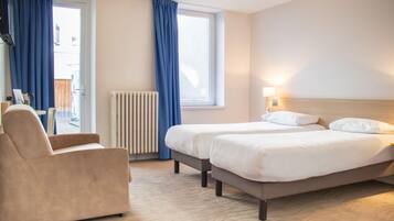 A hotel room with two beds, a chair, a window with curtains, a radiator, and a lamp.