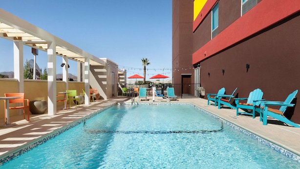 Una piscina con sillas de descanso y sombrillas, rodeada por un edificio de fachada roja y una pérgola blanca.