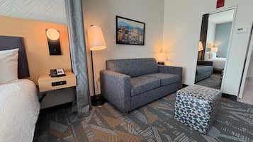 Una habitación de hotel con cama, sofá, escritorio con teléfono, lámpara y un cuadro de paisaje urbano en la pared.
