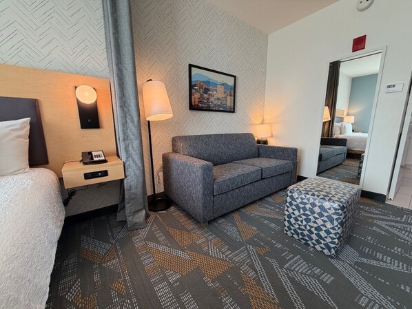 A hotel room with a bed, a sofa, a desk with a phone, a lamp, and a cityscape painting on the wall.
