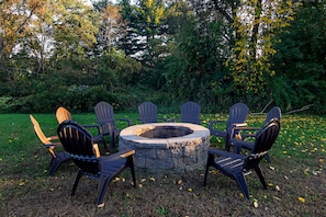 A circular stone fire pit surrounded by six Adirondack chairs in a garden setting. - Quality Inn Plainfield I-395 (Plainfield)