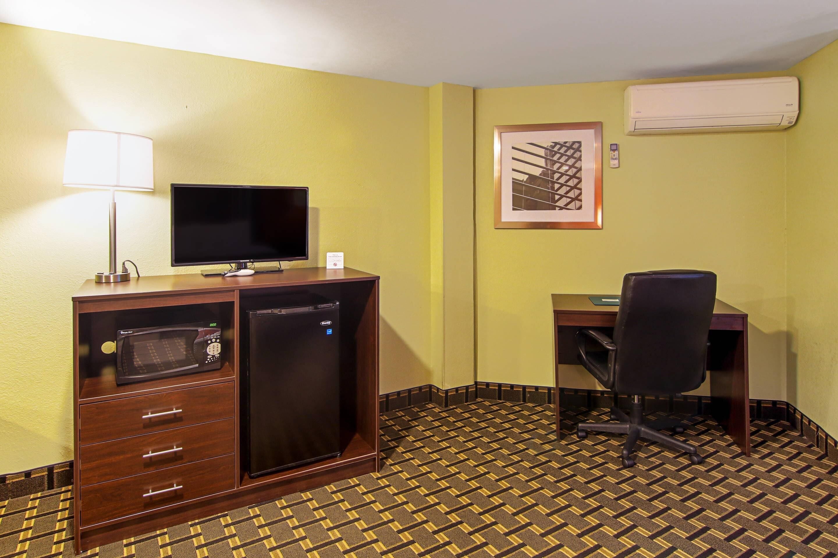 Hotelkamer met een bureau, een televisie, een magnetron, een koelkast, een stoel en een kamerbreed tapijt met dessin.