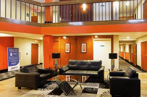 Un hall de hotel con una mesa de centro de vidrio, sofás de cuero, una escalera y un cartel.