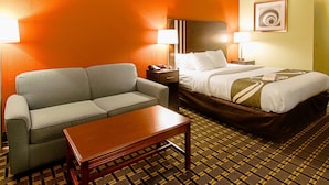 Una habitación de hotel con sofá, cama, mesita de noche, lámpara y alfombra estampada.