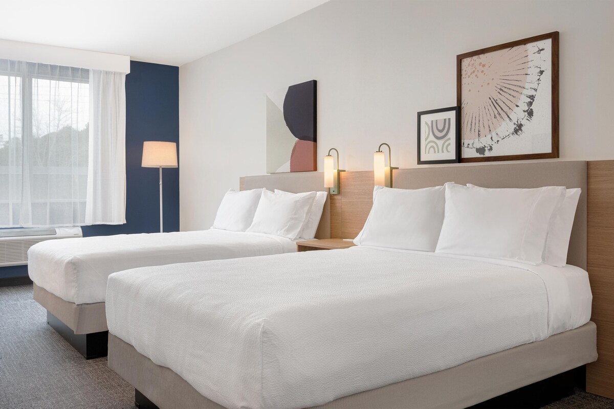 A hotel room with two beds, a window with sheer curtains, a standing lamp, and artwork on the walls.
