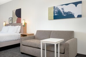A hotel room with a bed, a grey sofa, a small white table, and two wall-mounted lamps. - Spark by Hilton Douglasville Atlanta West (Douglasville)