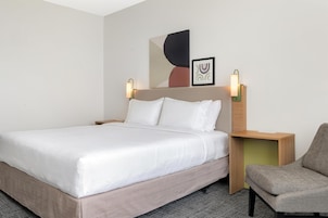 Habitación de hotel con una cama, una silla, dos lámparas fijadas a la pared y un cuadro abstracto enmarcado.