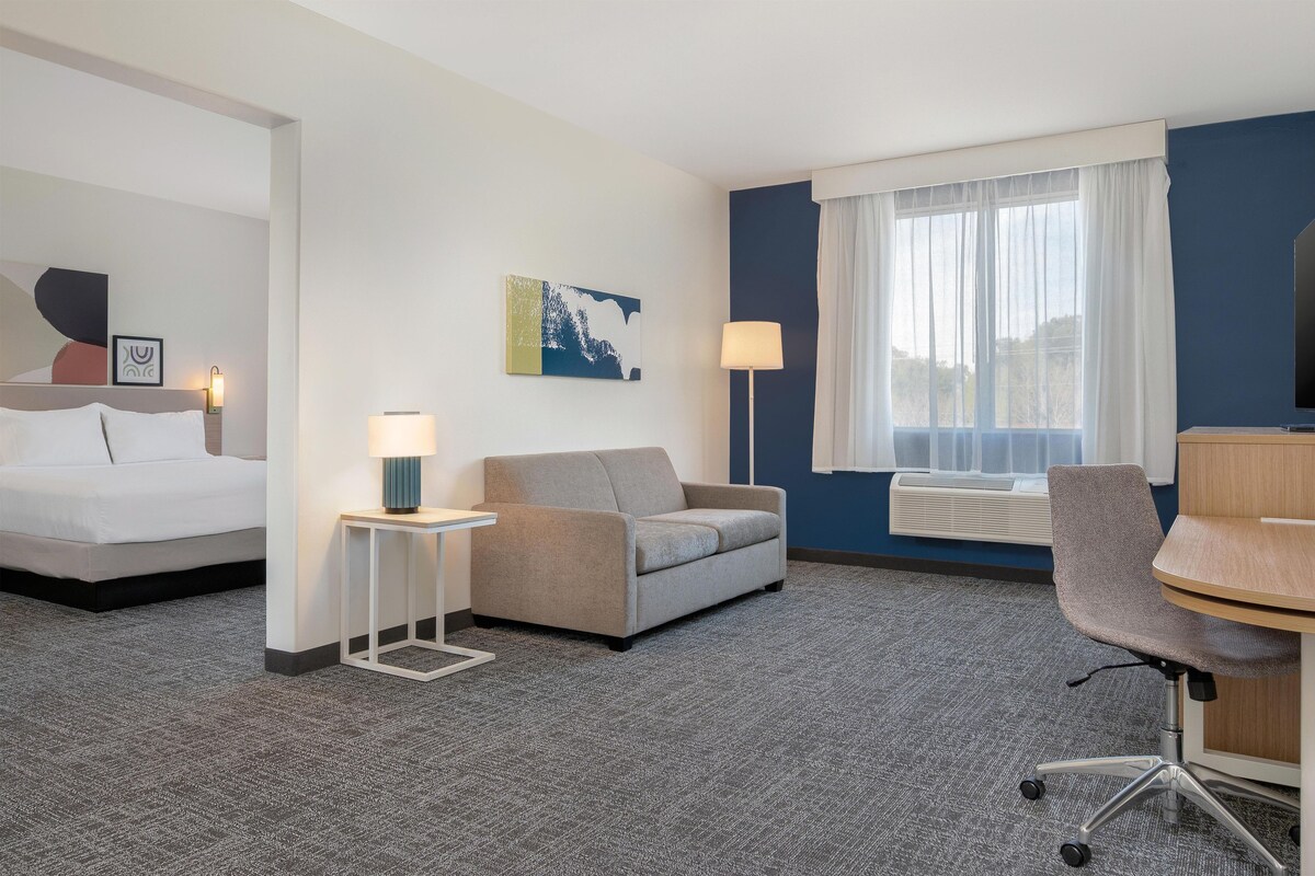 A hotel room with a bed, a sofa, a desk, and a chair. There is a window with curtains, a lamp, and a wall-mounted air conditioner.