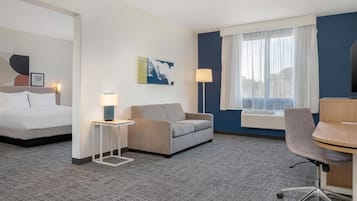 A hotel room with a bed, a sofa, a desk, and a chair. There is a window with curtains, a lamp, and a wall-mounted air conditioner.