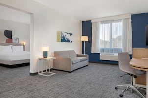 Habitación de hotel con cama, sofá, escritorio y silla. Hay una ventana con cortinas, una lámpara y un aire acondicionado instalado en la pared.