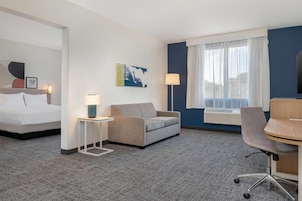 A hotel room with a bed, a sofa, a desk, a chair, a lamp, a painting, and a window with curtains.