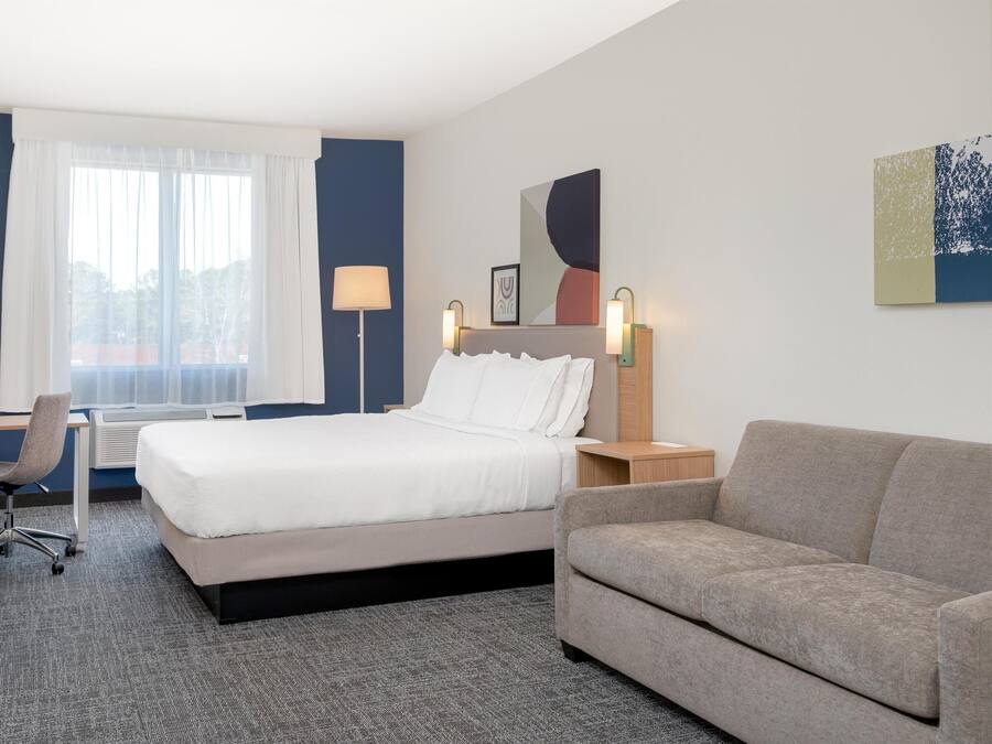 Une chambre d’hôtel avec un lit, un bureau, une chaise, un canapé et une grande fenêtre avec des rideaux.