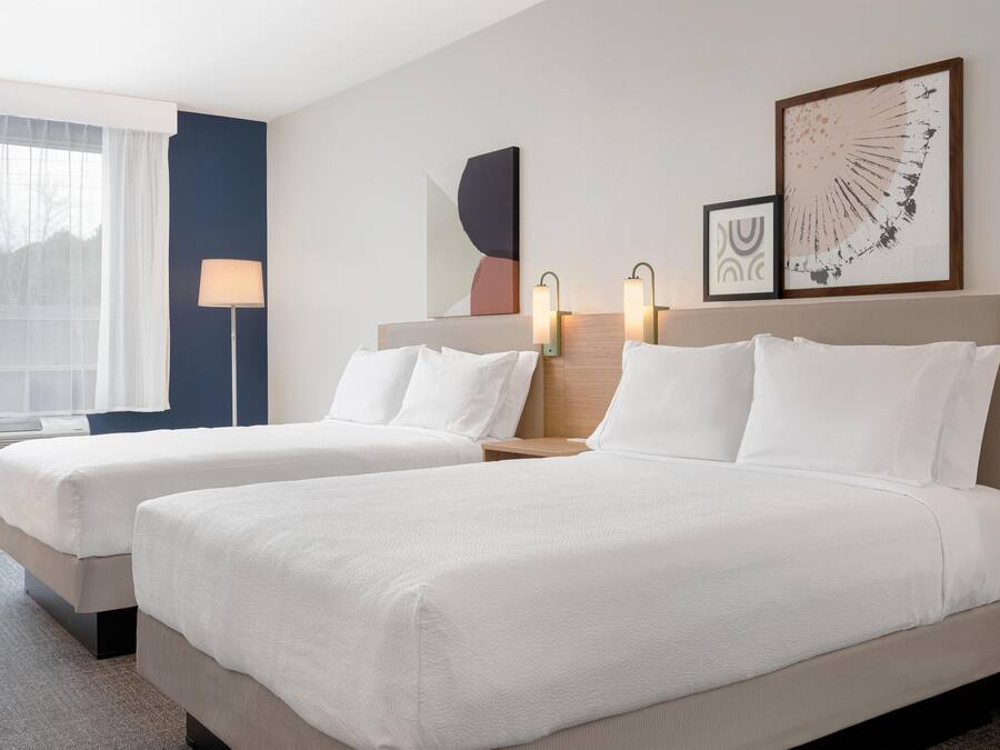 Une chambre d’hôtel avec deux lits, une fenêtre avec des rideaux légers, une lampe sur pied et des œuvres d’art accrochées au mur.