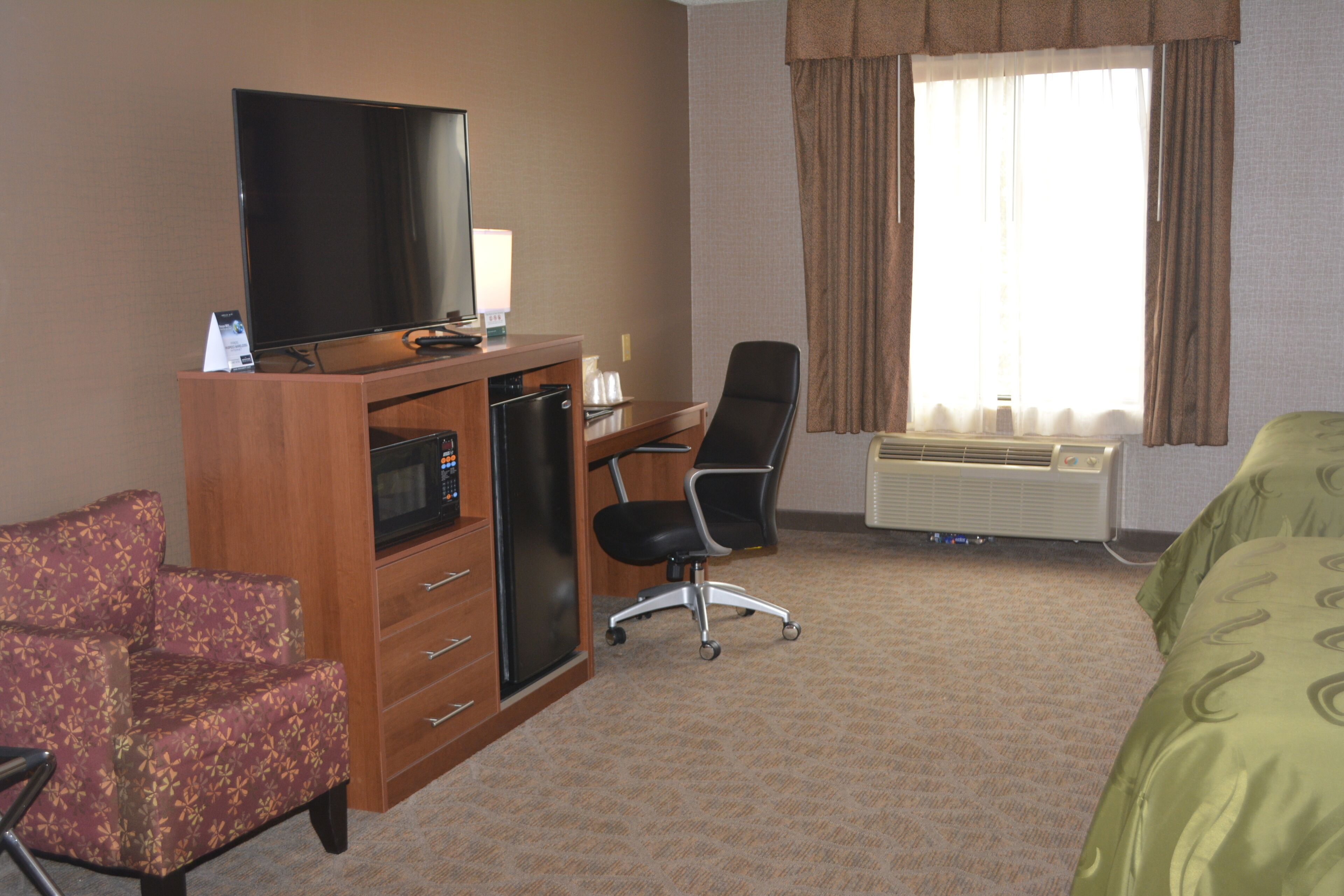 A hotel room with a television on a wooden cabinet, a desk with a chair, a bed with a green bedspread, and an air conditioning unit.