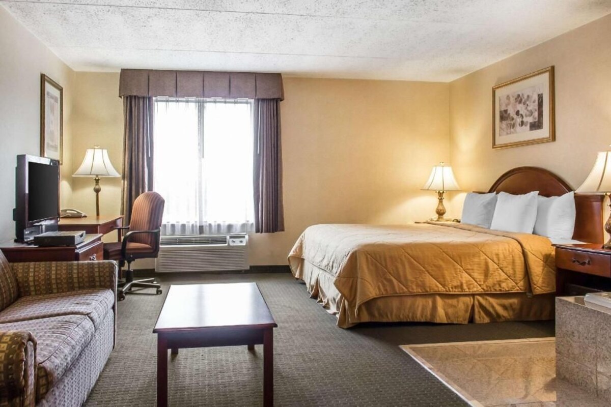 Habitación de hotel con cama, sofá, escritorio con televisor, lámpara y ventana con cortinas.
