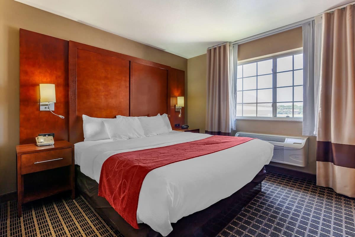 A hotel room with a large bed, a wooden headboard, two bedside lamps, a nightstand, a window with curtains, and an air conditioning unit.