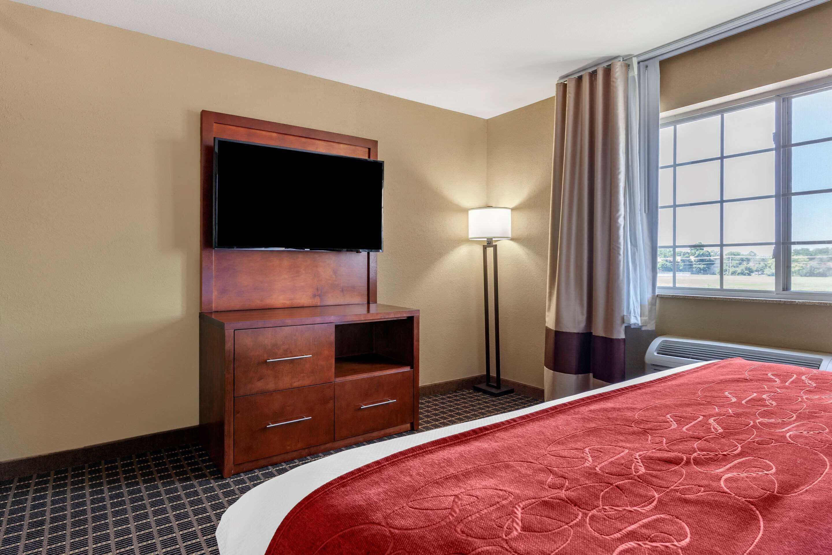 Une chambre d’hôtel avec un lit, un téléviseur fixé à un meuble en bois, une lampe sur pied et une fenêtre avec des rideaux.