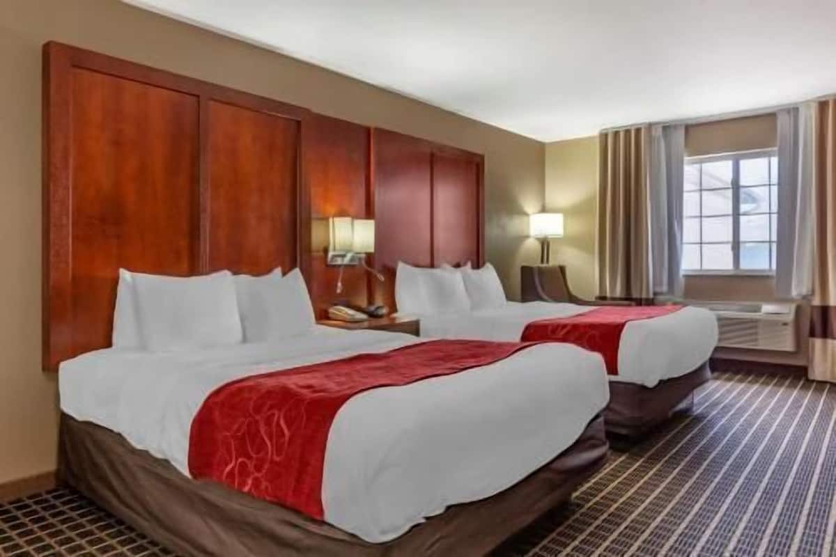 A hotel room with two beds, a large wooden headboard, a window with curtains, and a patterned carpet.