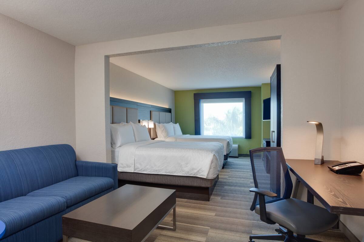 Una habitación de hotel con un sofá azul, un escritorio de madera, una silla, dos camas y un ventanal grande.