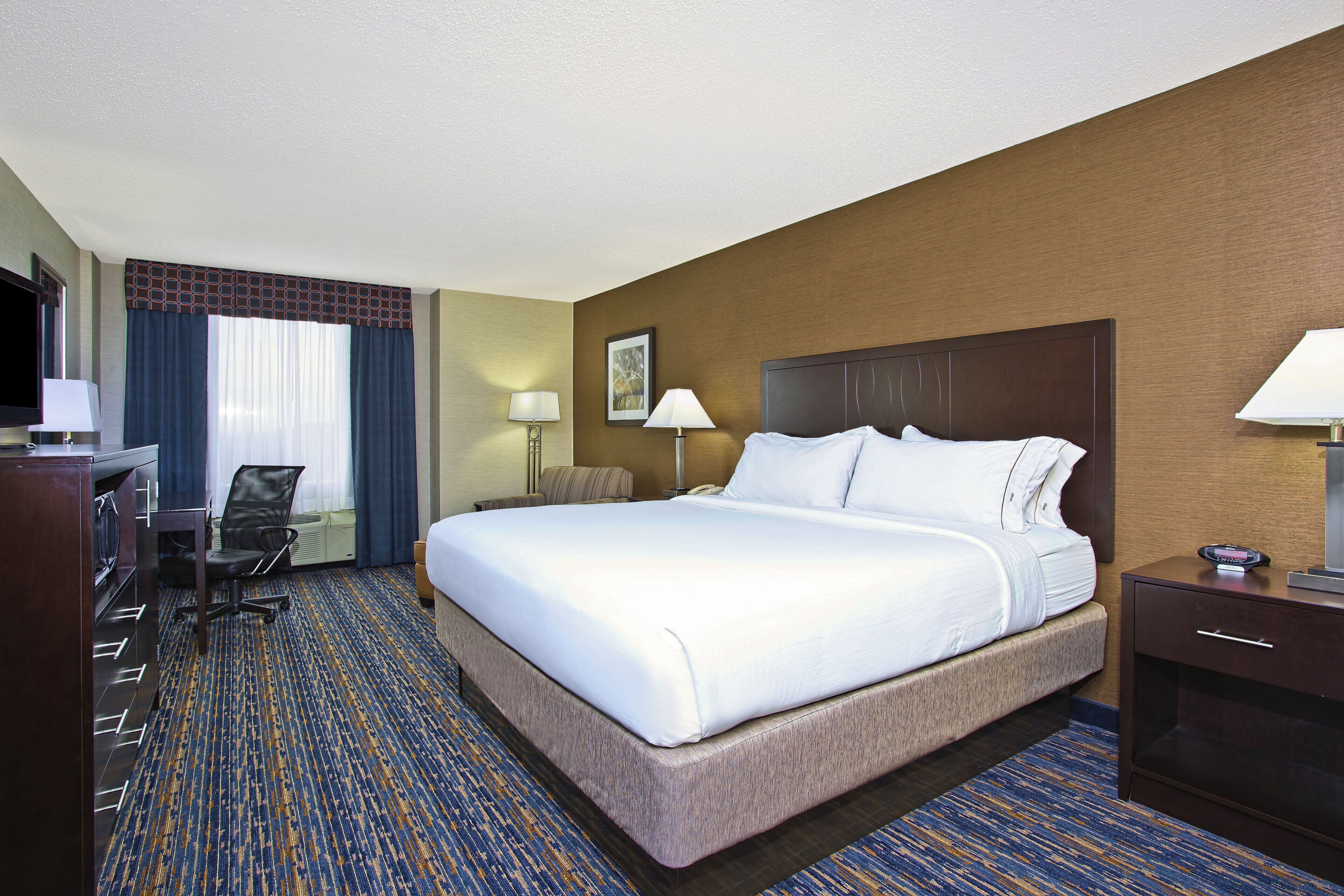 A hotel room with a large bed, a desk with a chair, a TV, and a lamp.