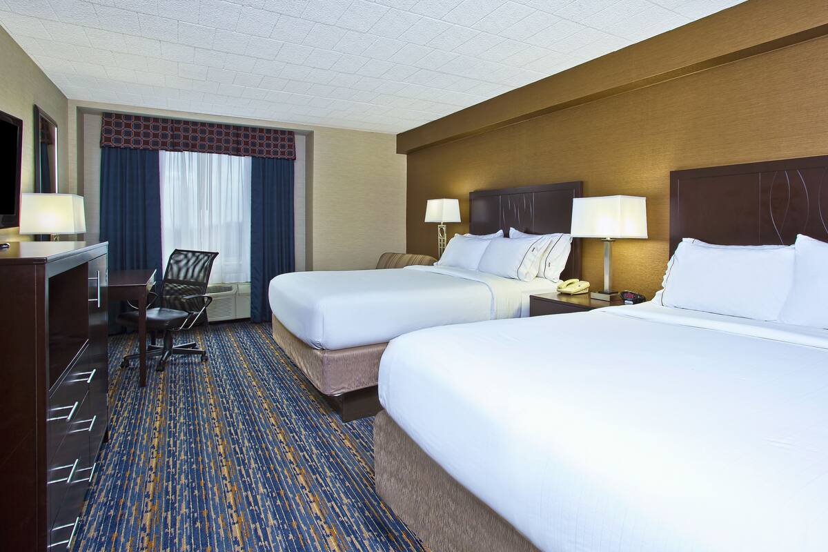 A hotel room with two beds, a desk, a chair, and a nightstand with a lamp.