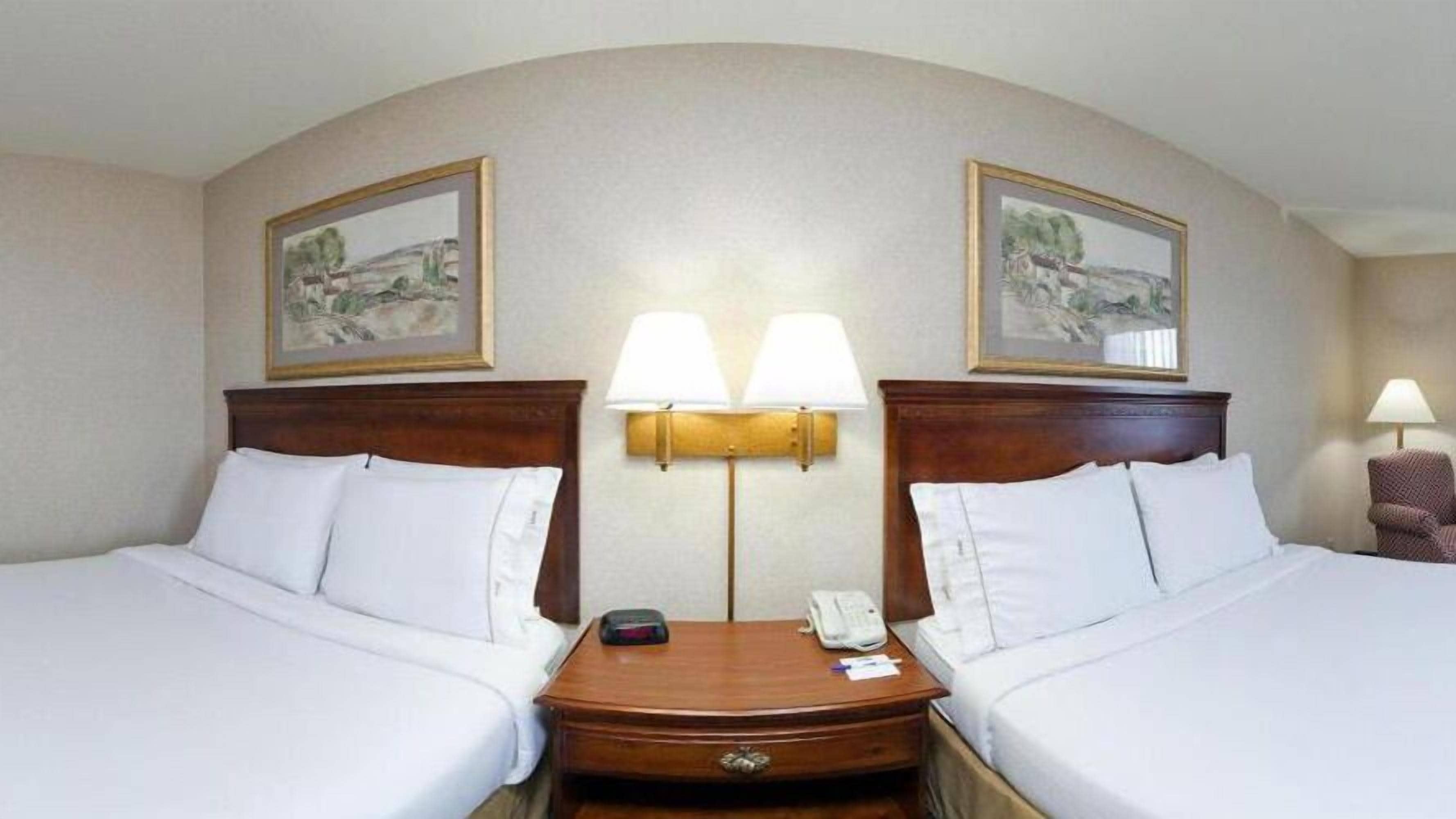 Hotellihuone, jossa on kaksi sänkyä, yöpöytä, lamppu ja kaksi kehystettyä kuvaa seinällä.