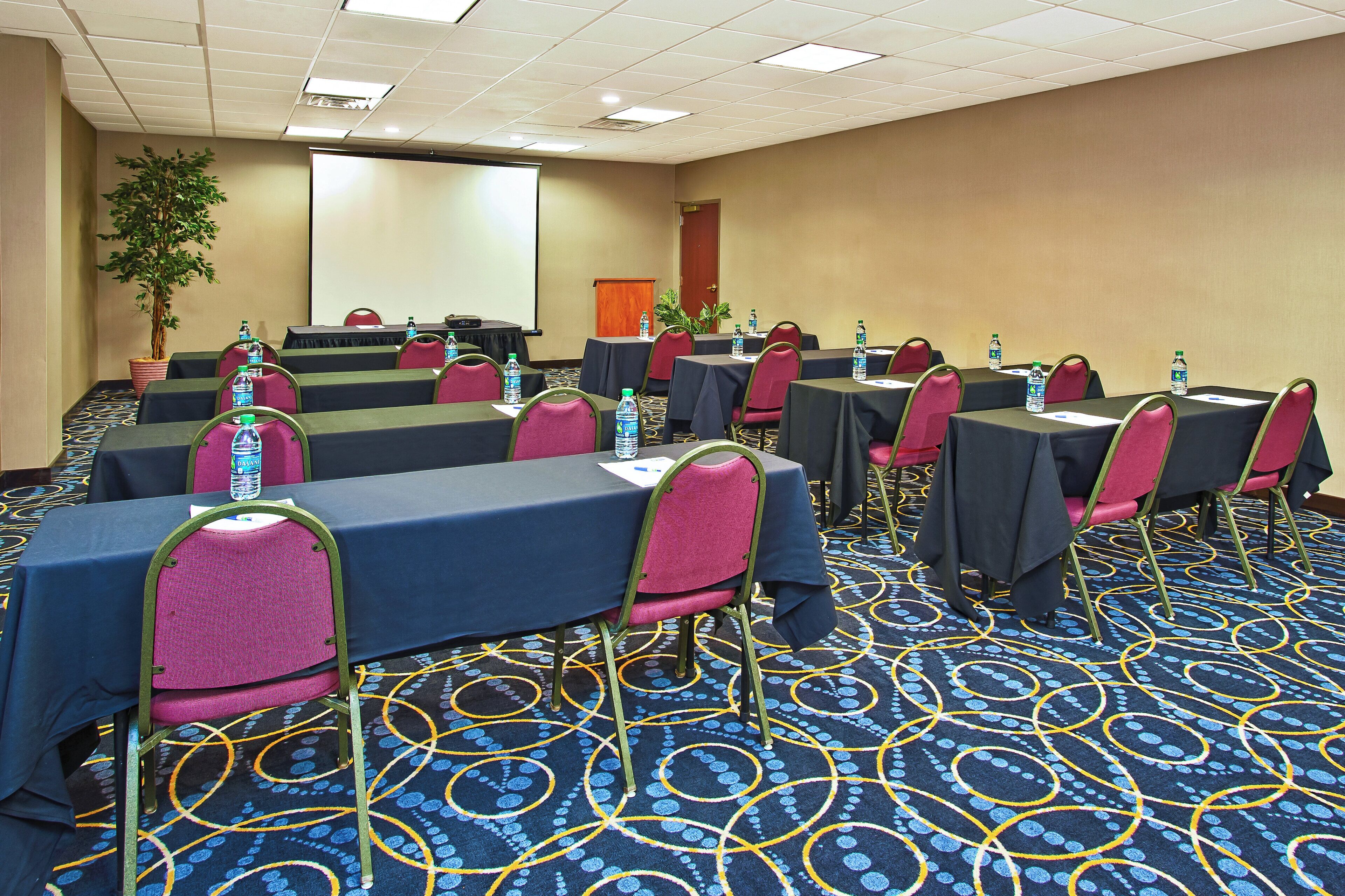 A conference room with rows of tables and chairs, water bottles on each table, and a projector screen at the front.