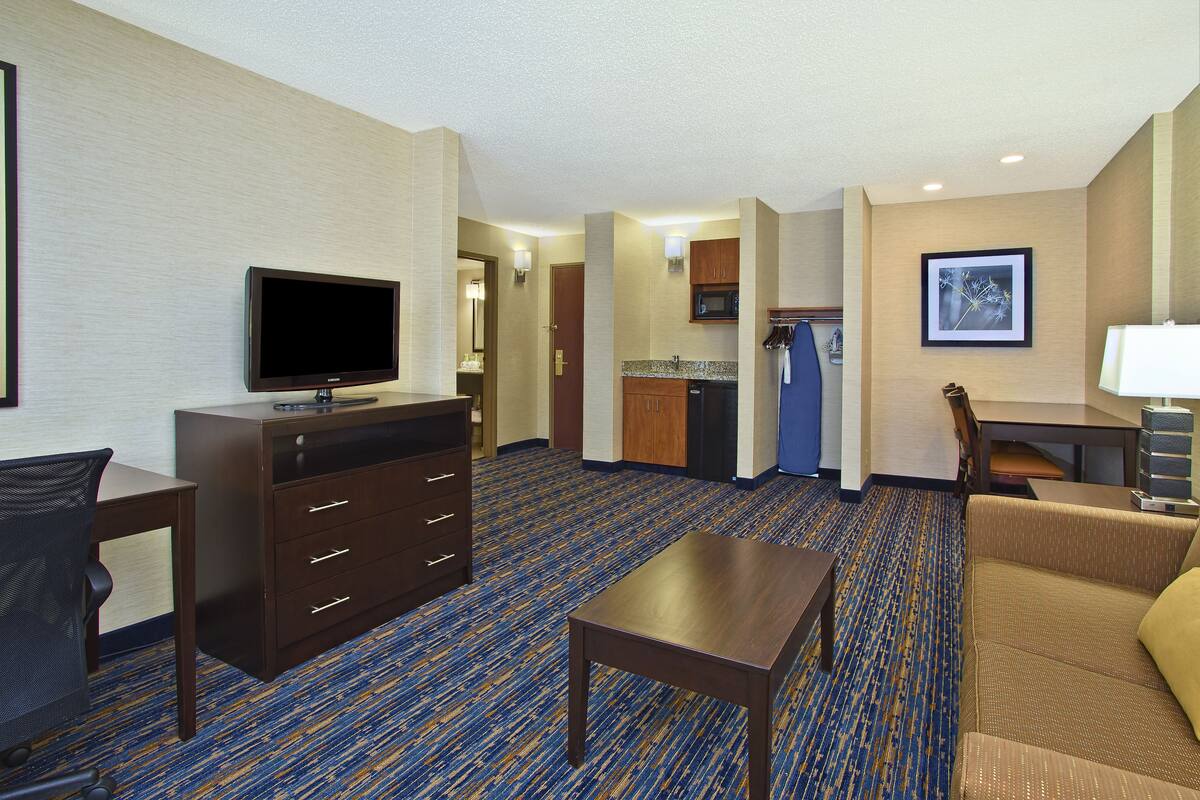 A hotel room with a flat-screen TV, a desk, a sofa, a dresser, and a kitchenette.