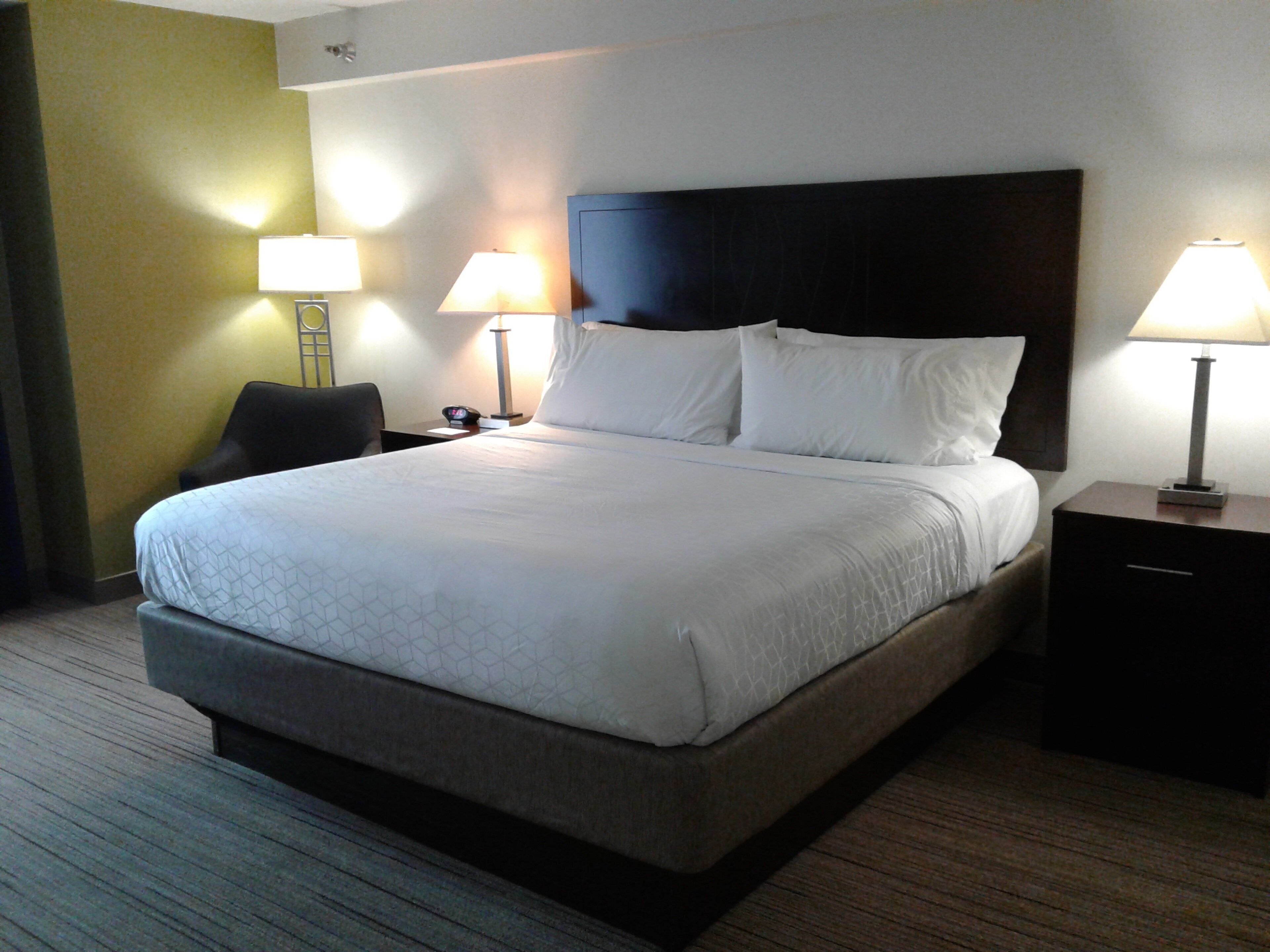A hotel room with a large bed, two bedside tables with lamps, a chair, and a nightstand with a phone.