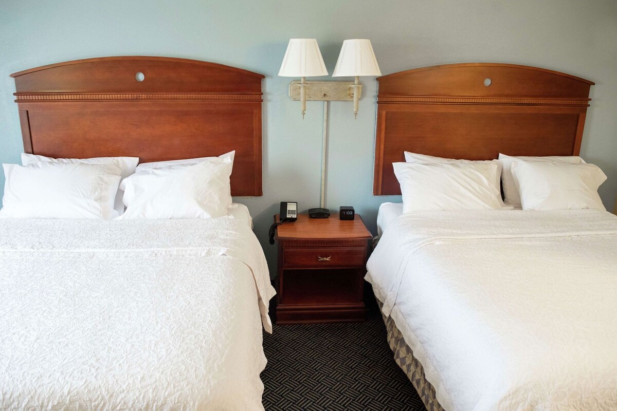 A hotel room with two beds, a nightstand with a phone and clock, and two wall-mounted lamps.