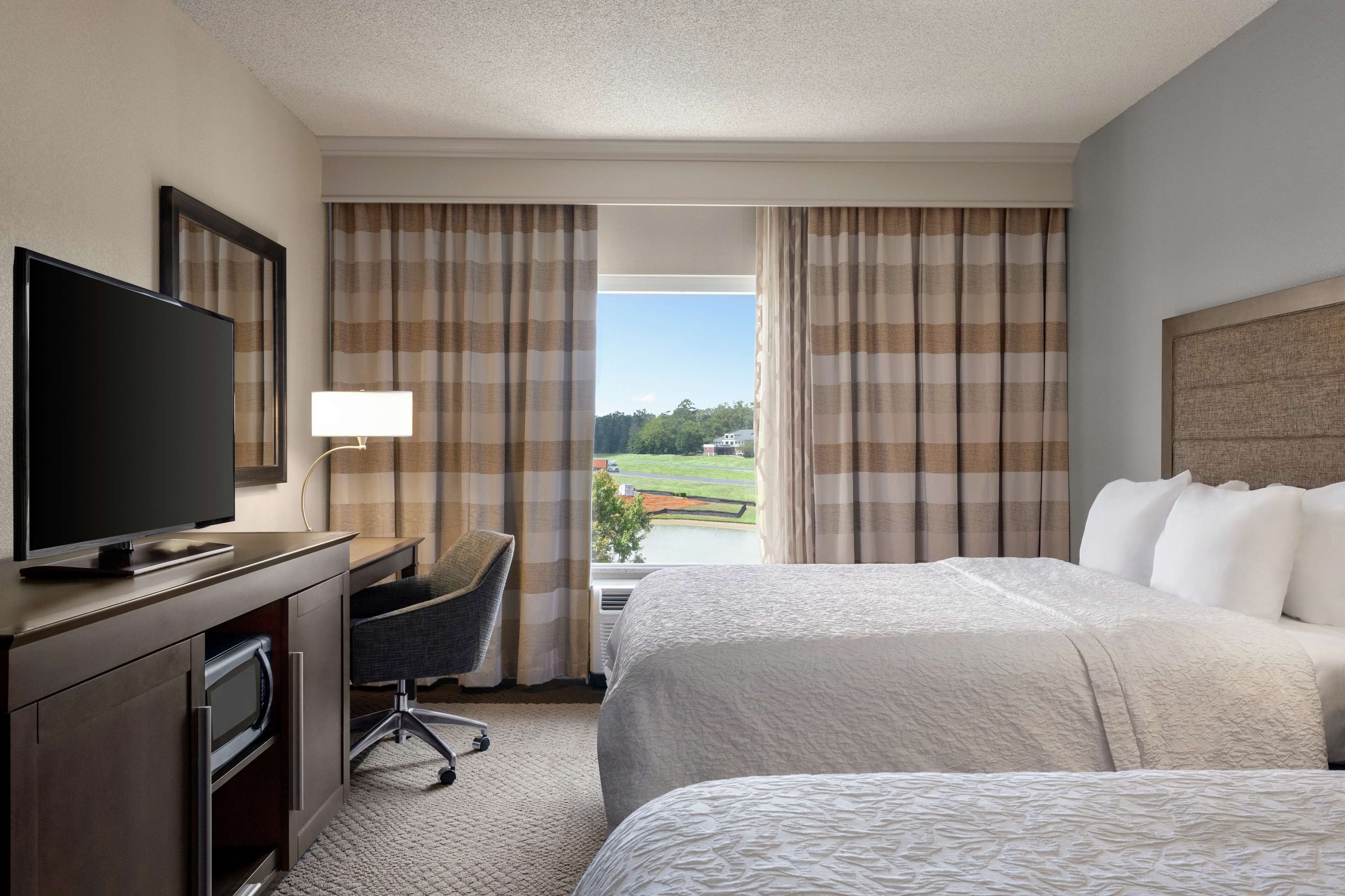 Une chambre d’hôtel avec deux lits, une télévision, un bureau avec une chaise et une vue sur un paysage verdoyant, visible à travers les rideaux.