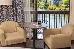 A room with two beige armchairs, a round wooden table with a book and a mug, a view of a river through a balcony railing.