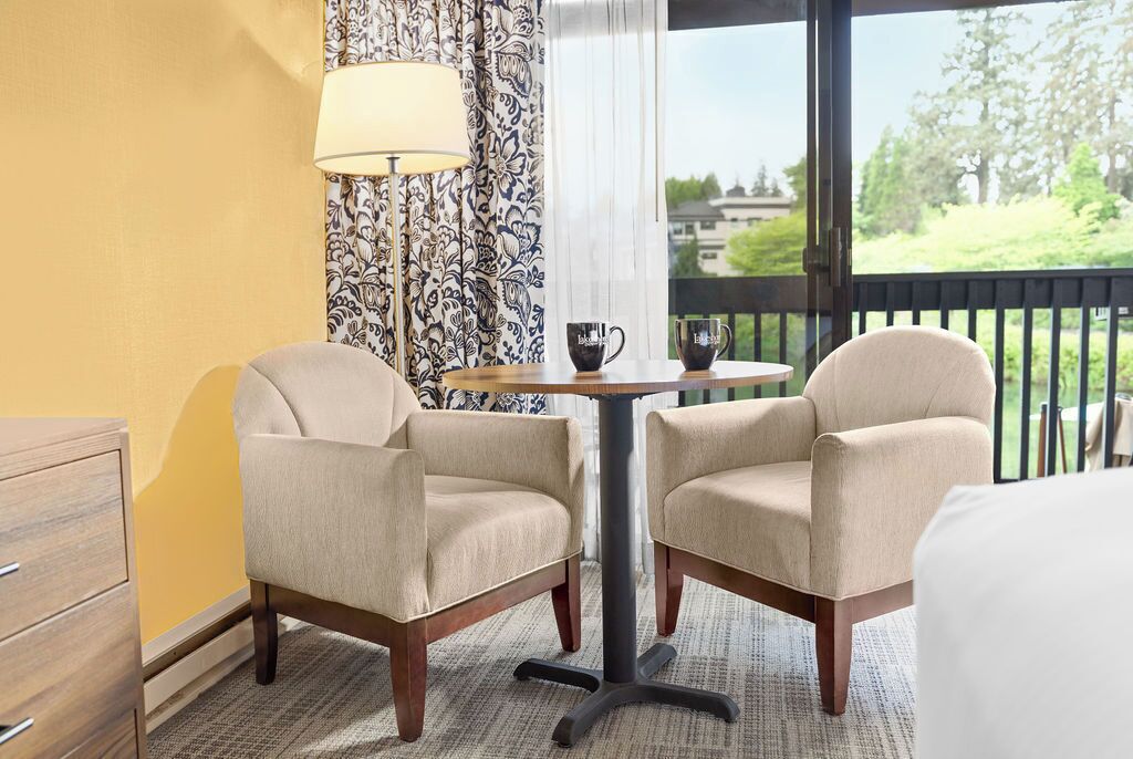 A hotel room with two armchairs, a small round table with a lamp, and a view of a balcony.